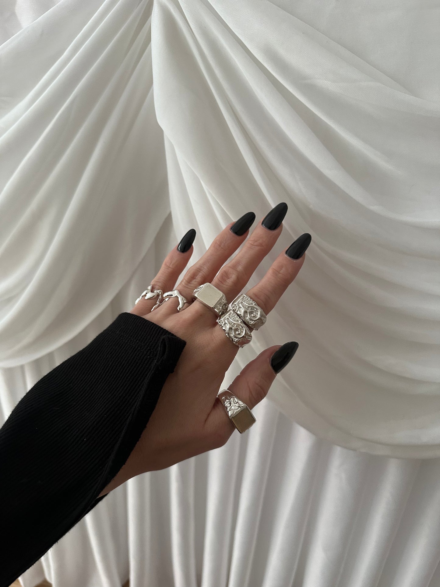 Hand with multiple rings and black nail polish against a white draped fabric background